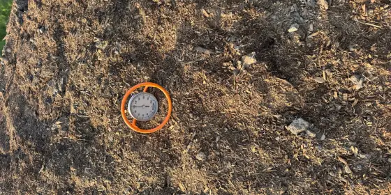 Compost thermometer in an orange guard lying on a Casco Compost windrow, dark brown wood chips and soil around it