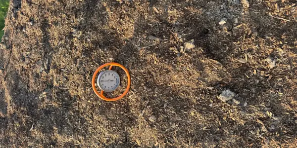 Compost thermometer in an orange guard lying on a Casco Compost windrow, dark brown wood chips and soil around it