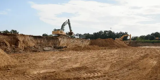Excavadora trabajando al borde de una fosa de arena, con huellas de llantas al frente y cielo azul