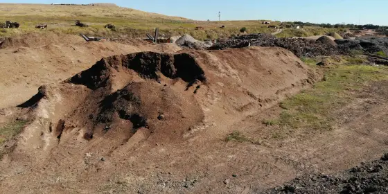 Largas hileras de madera molida y material orgánico café, con una loma de pasto y maquinaria atrás