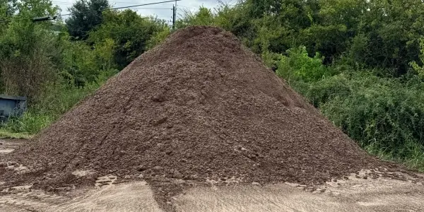 Pile of brown Lawn Mix on sandy ground in the yard, with trees and brush behind