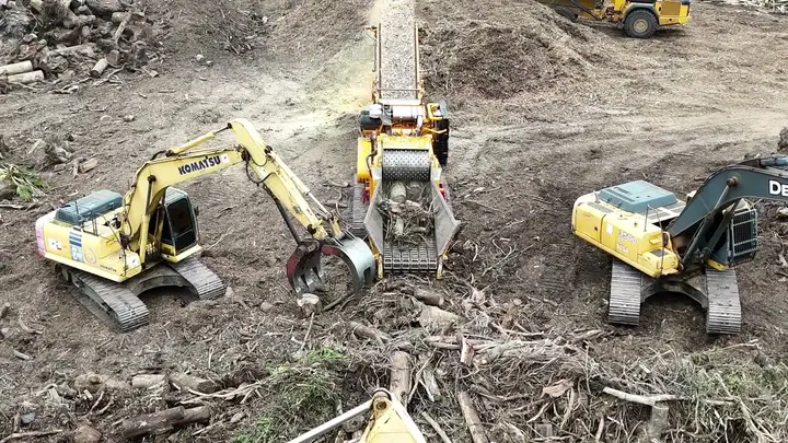 An excavator with a grapple feeds stumps and brush into a grinder while ground wood comes off the discharge conveyor and a second excavator stands by