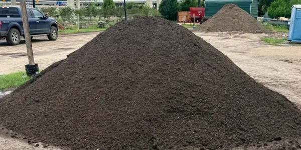 Pile of dark brown Tree & Shrub Mix in the retail yard, with a second pile, a pickup truck and the freeway overpass behind
