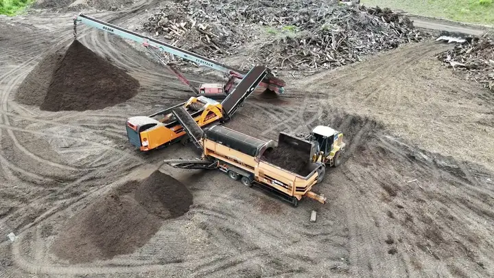 Aerial view of a wheel loader tipping a bucket of dark material into the hopper of a long orange trommel, with a conveyor stacking a tall dark pile and a lower pile beside the drum